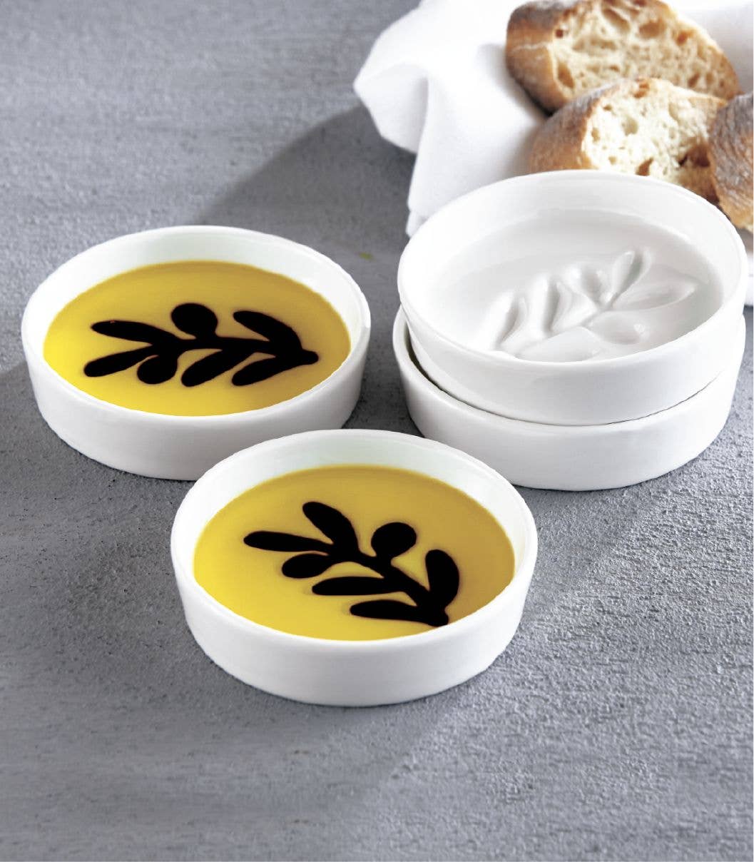 Jeanne Fitz white dipping plates with olive oil and balsamic vinegar in an olive branch pattern, alongside sliced bread.