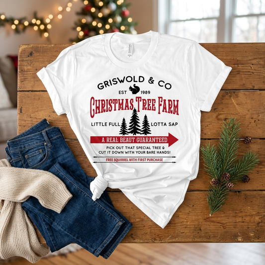 White Griswold Christmas Tree Farm graphic T-shirt on a wooden bench beside jeans and a knitted scarf with Christmas lights behind